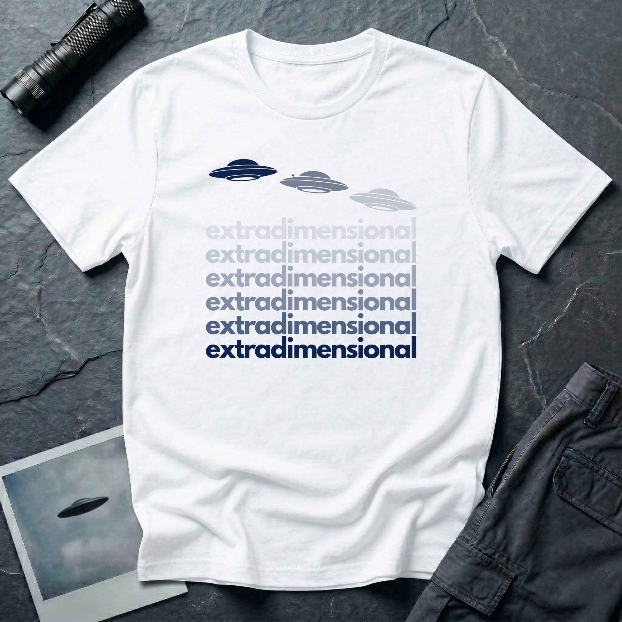 White t-shirt with three UFO saucer silhouettes in navy to grey gradient above the word extradimensional repeated six times in stacked bold type fading from grey to navy