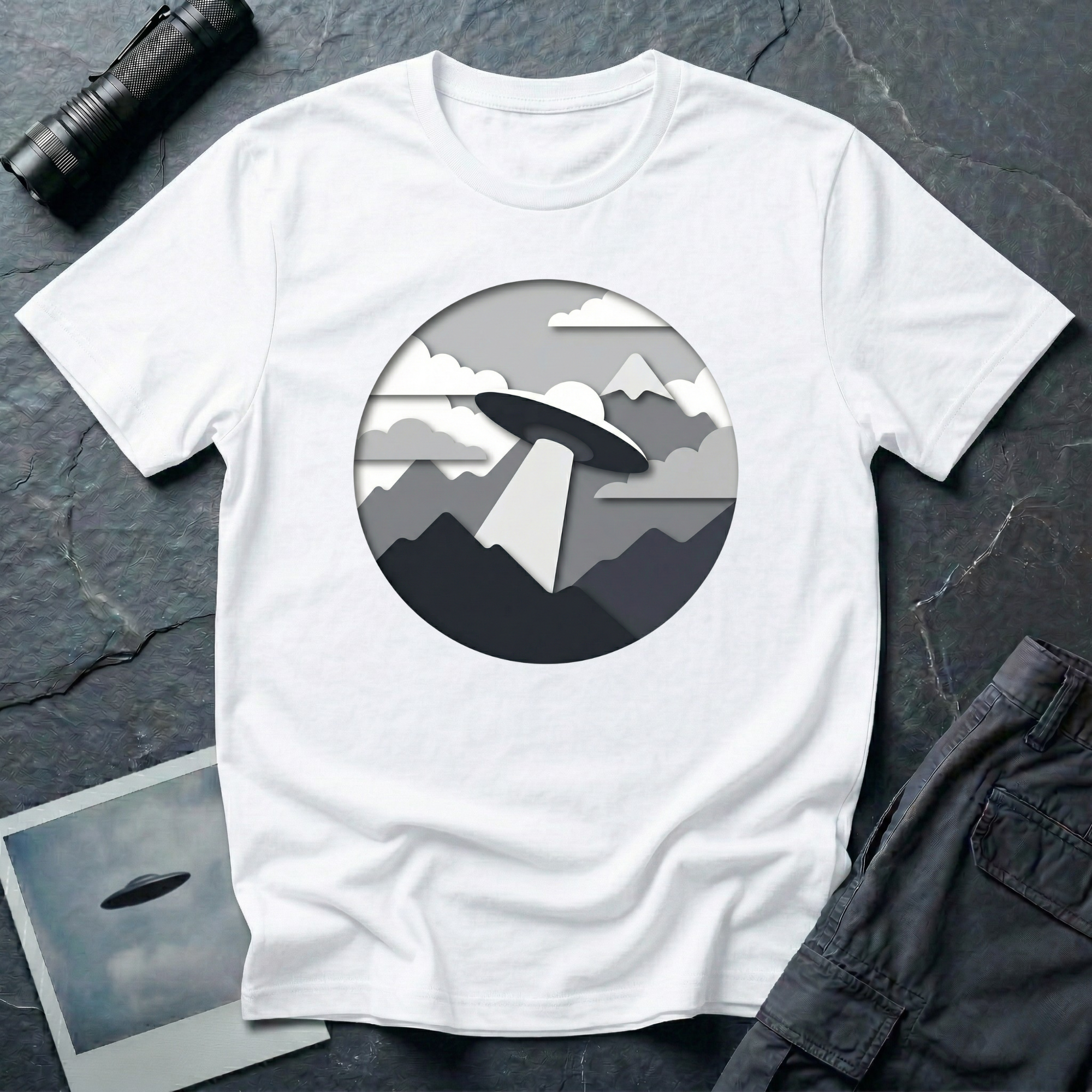 Papercut UFO t-shirt — layered paper-craft illustration of flying saucer over mountains and clouds in grey monochrome on white cotton tee