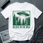 Believe In The Wild forest UFO t-shirt — flying saucer over dense pine forest rendered in green woodcut linocut carved-block print style with "BELIEVE IN THE WILD" caption on white cotton tee