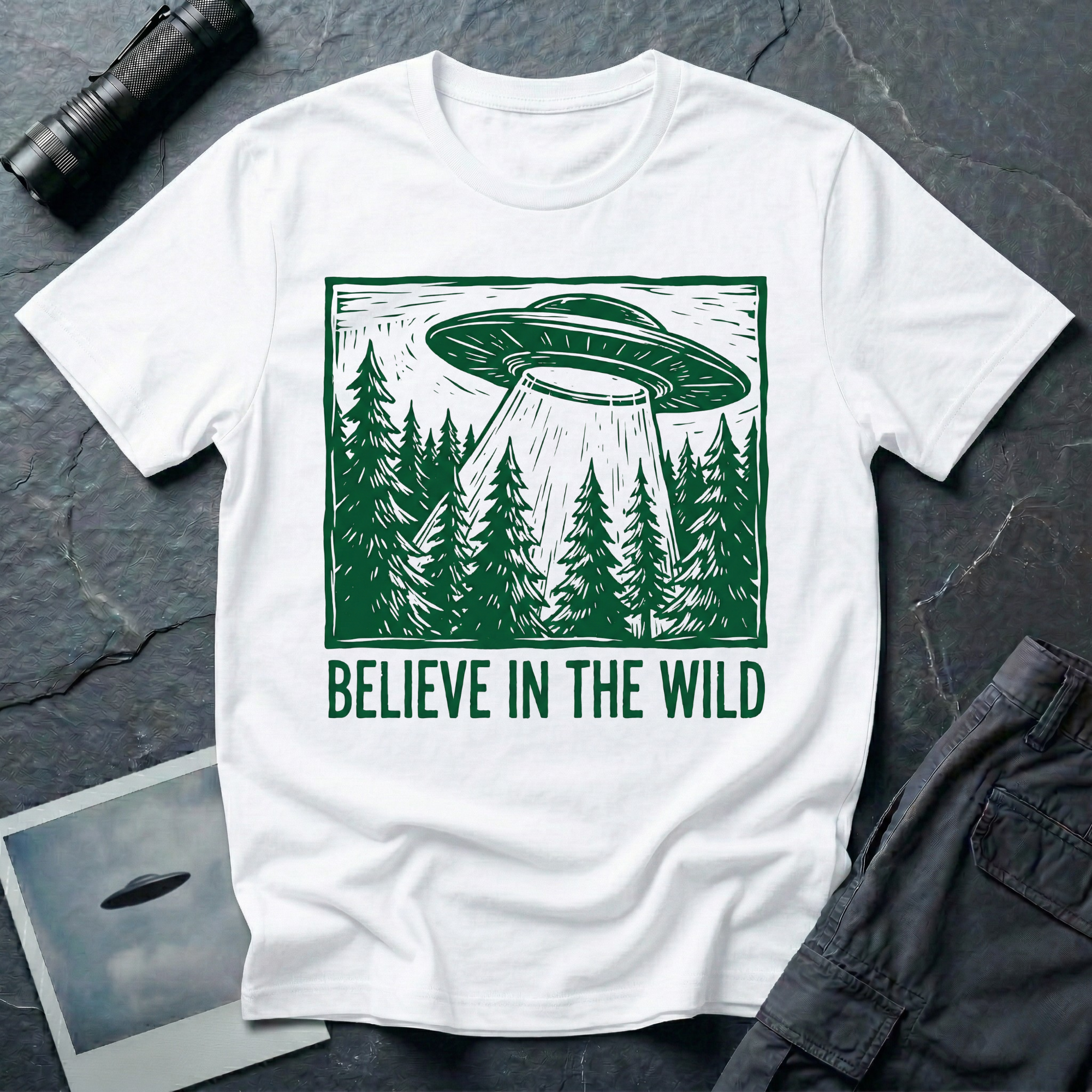 Believe In The Wild forest UFO t-shirt — flying saucer over dense pine forest rendered in green woodcut linocut carved-block print style with "BELIEVE IN THE WILD" caption on white cotton tee