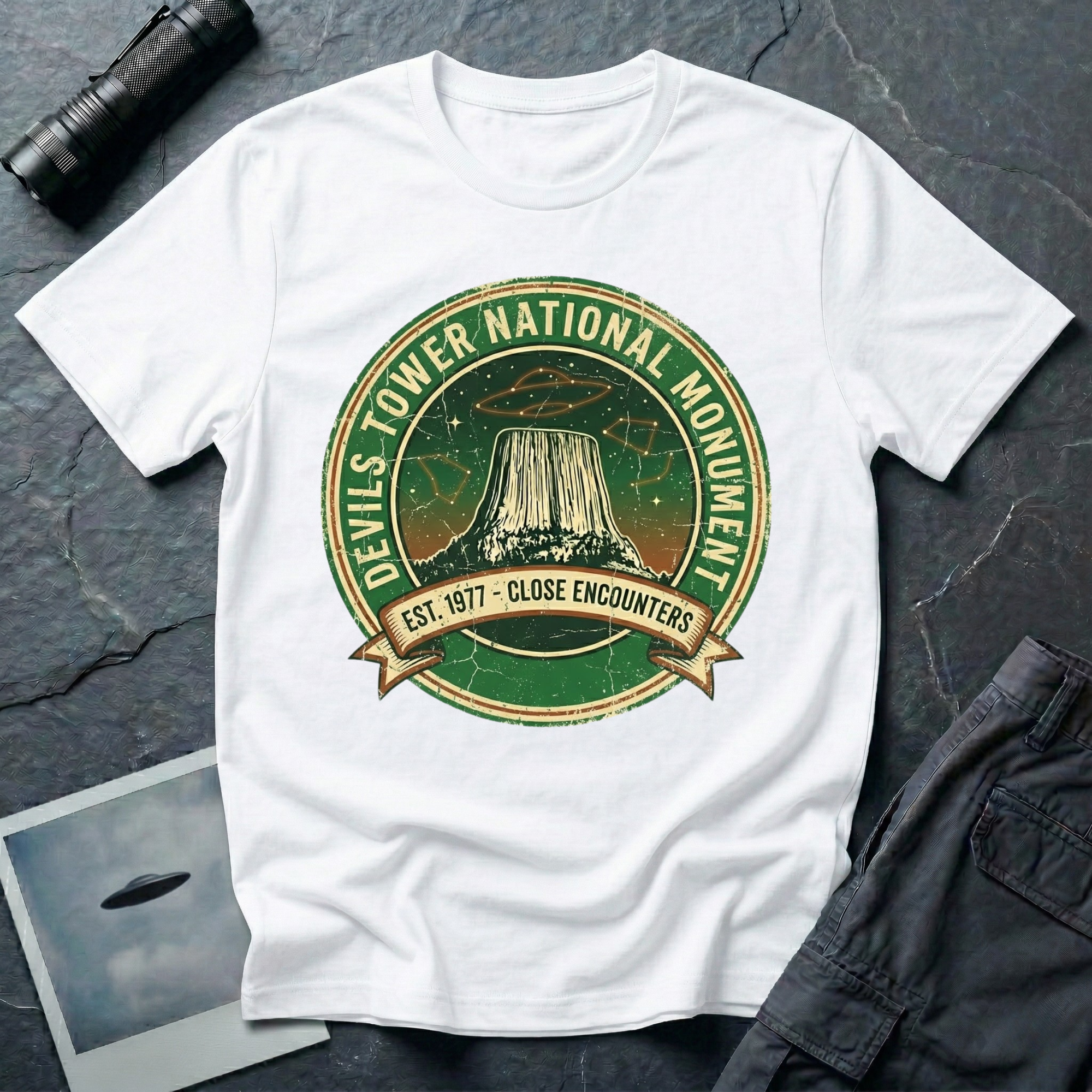 Devils Tower UFO t-shirt — vintage distressed National Park badge with the Wyoming monument silhouette, tiny flying saucers and constellations over a starry green sky, "DEVILS TOWER NATIONAL MONUMENT" arc text and "EST. 1906 — CLOSE ENCOUNTERS" banner on white cotton tee