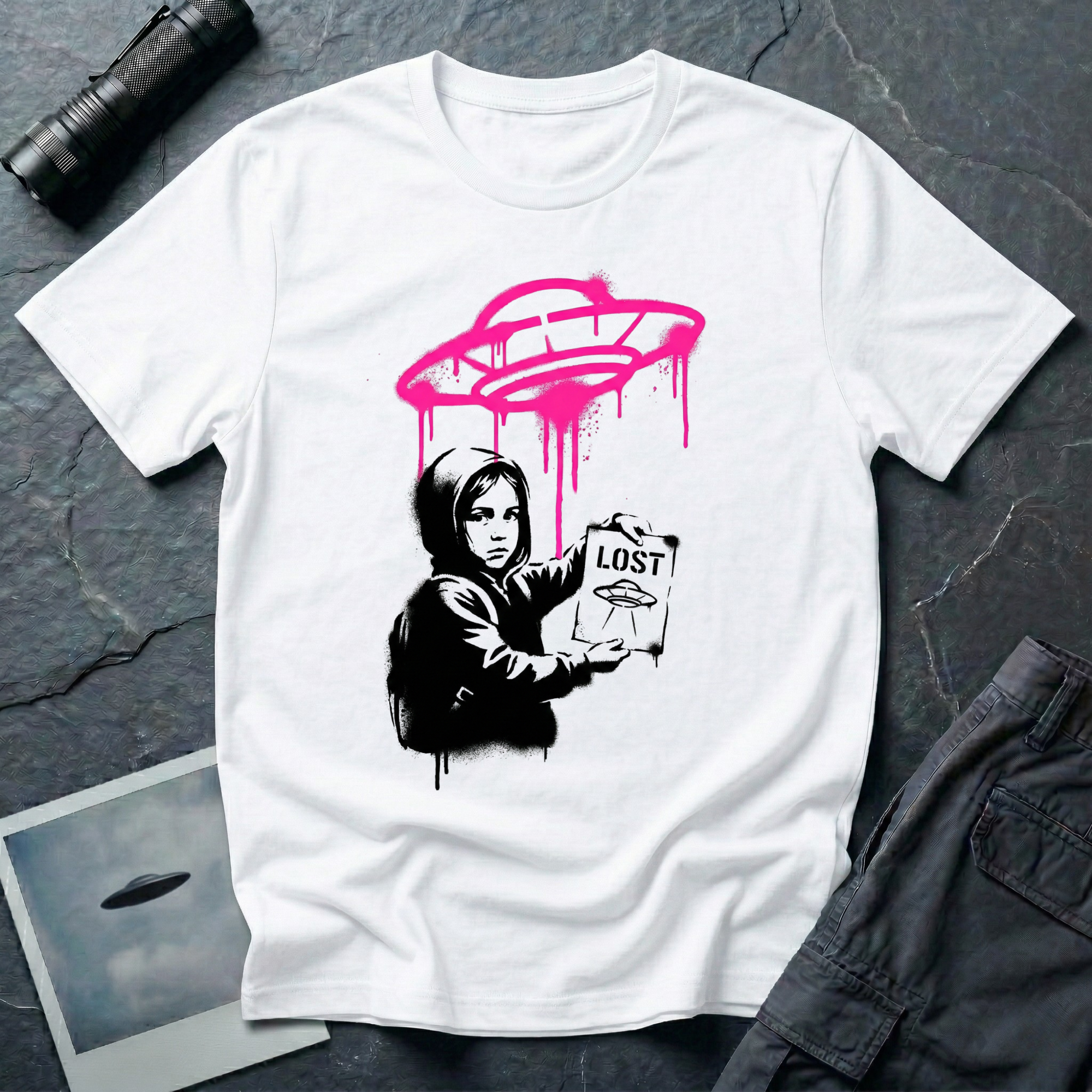 Street art UFO t-shirt with black stencil hooded figure holding "LOST" poster featuring flying saucer icon, bright magenta spray-painted UFO with paint drips hovering above in Banksy graffiti style on white cotton tee