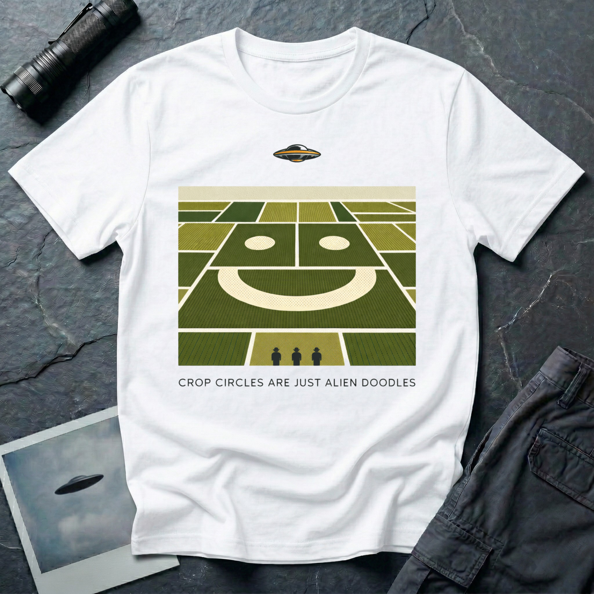 Alien Doodles crop circle humor t-shirt — aerial view of farmland fields forming a giant smiley face with tiny flying saucer hovering above and three small human figures below, "CROP CIRCLES ARE JUST ALIEN DOODLES" caption on white cotton tee