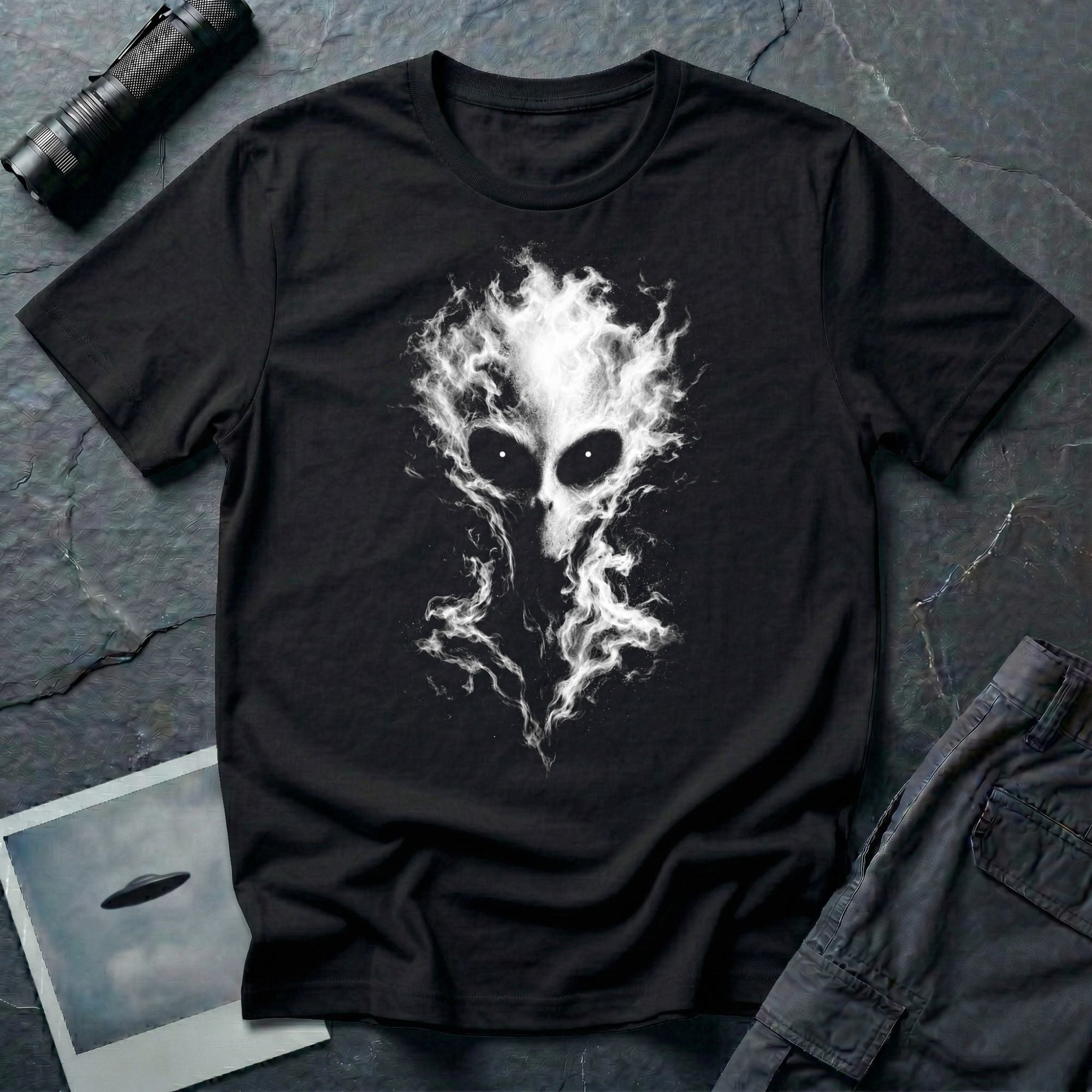 Alien t-shirt laid flat on a dark textured surface, featuring a large white and gray alien head made of smoke, with glowing eyes, black