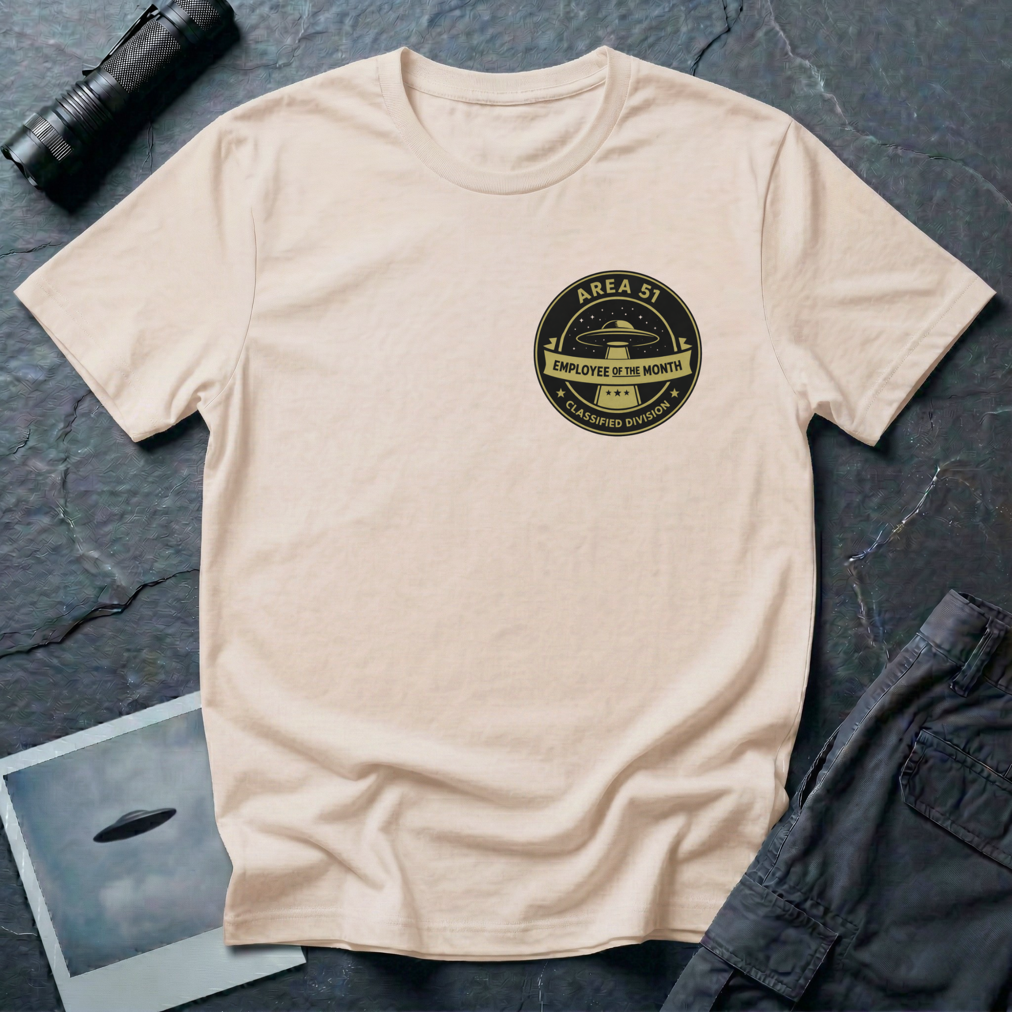 Area 51 Employee Of The Month UFO t-shirt — vintage circular badge with classic flying saucer, "Area 51" and "Classified Division" arc text, "Employee of the Month" ribbon banner in olive and black on upper-left chest of natural cotton tee