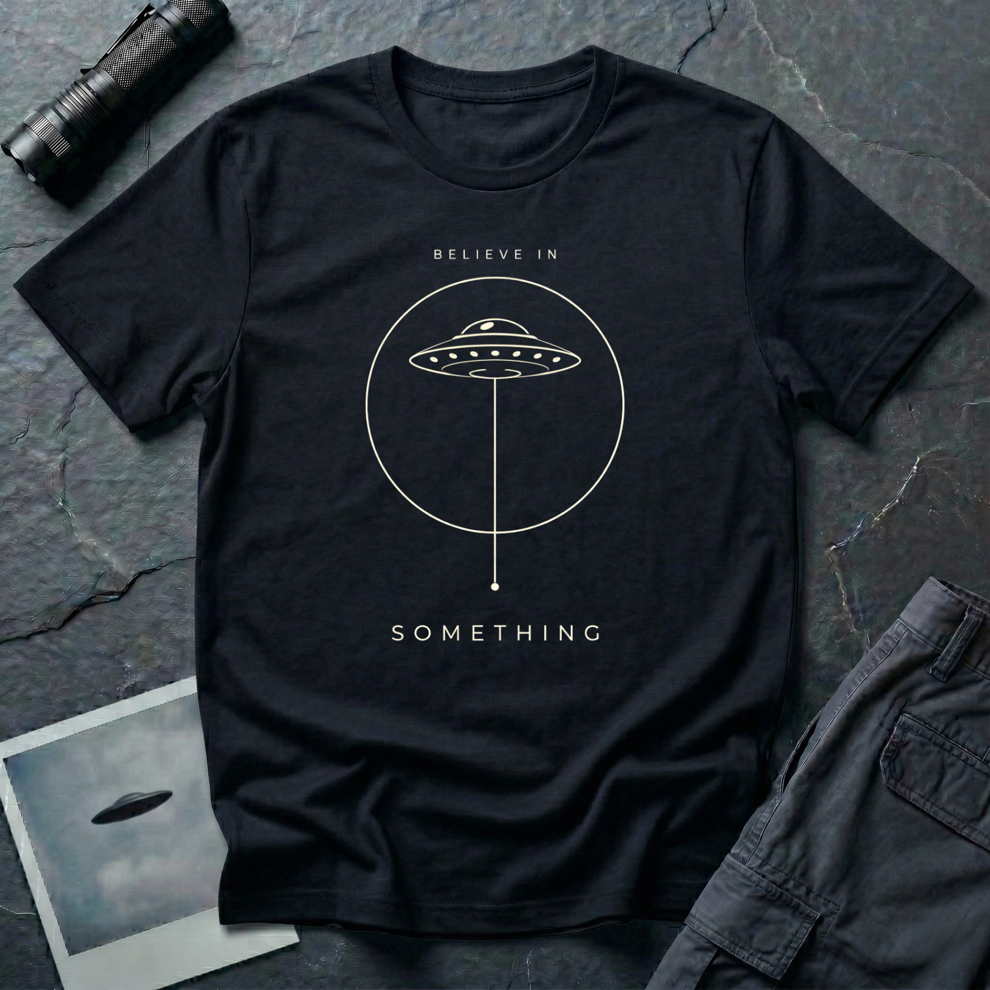 Believe in Something minimalist UFO t-shirt — flying saucer inside a circle with a suspended plumb line in cream on black cotton tee