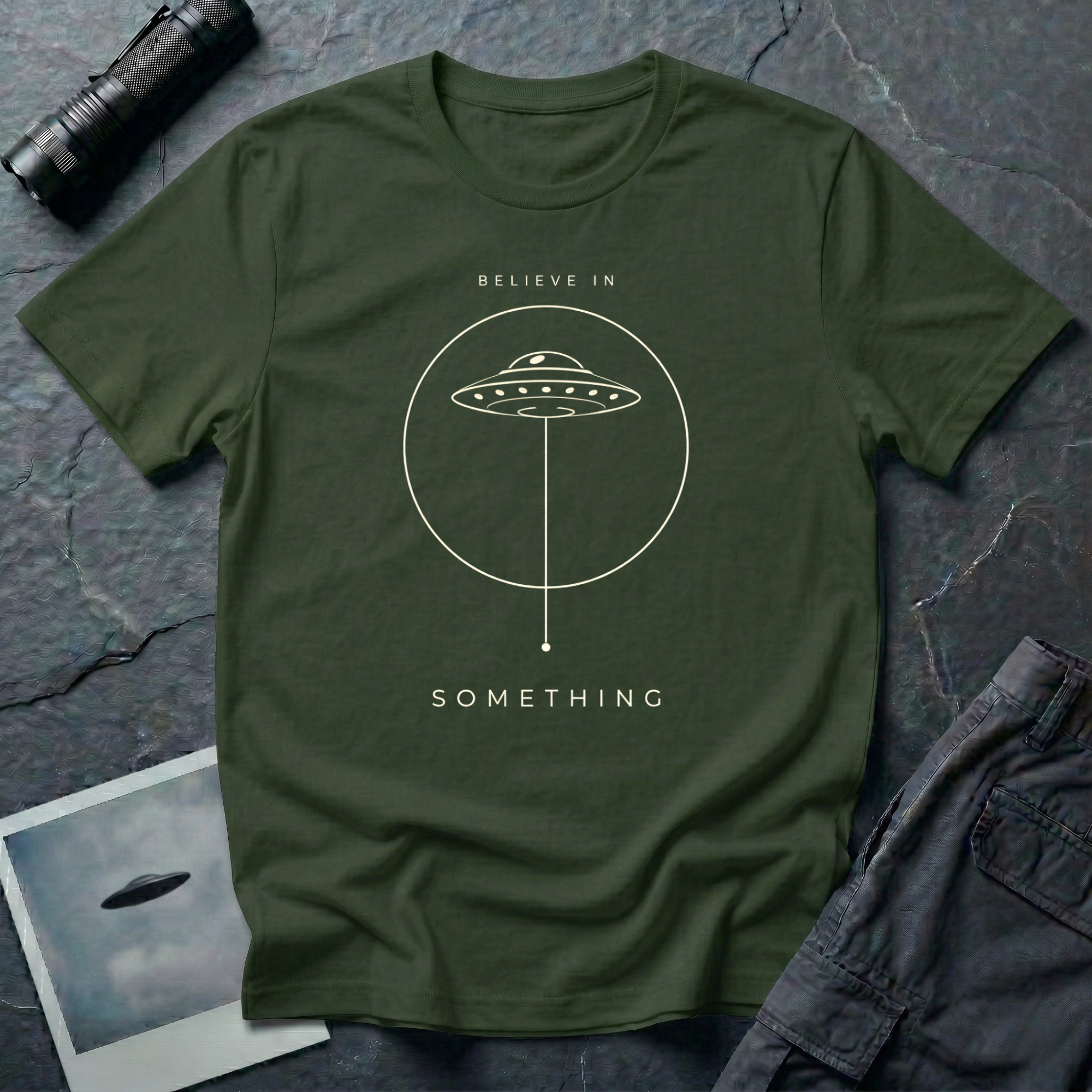 Believe in Something minimalist UFO t-shirt — flying saucer inside a circle with a suspended plumb line in cream on military green cotton tee
