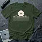 Desert Crossing UFO T-shirt with a black flying saucer crossing a white moon above minimalist line-art desert dunes, military green tee