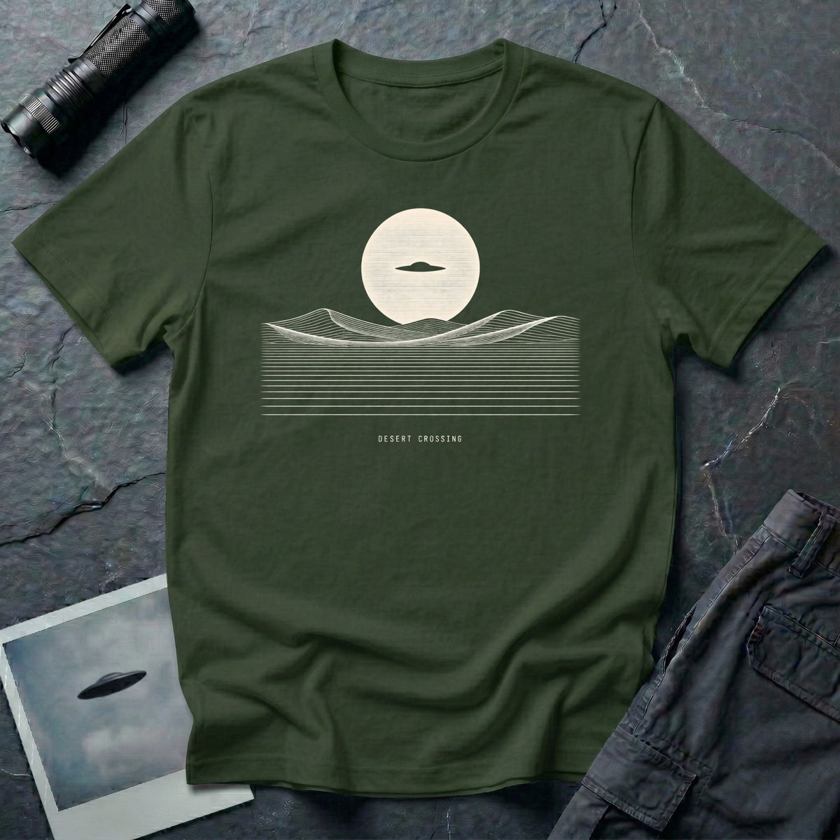 Desert Crossing UFO T-shirt with a black flying saucer crossing a white moon above minimalist line-art desert dunes, military green tee