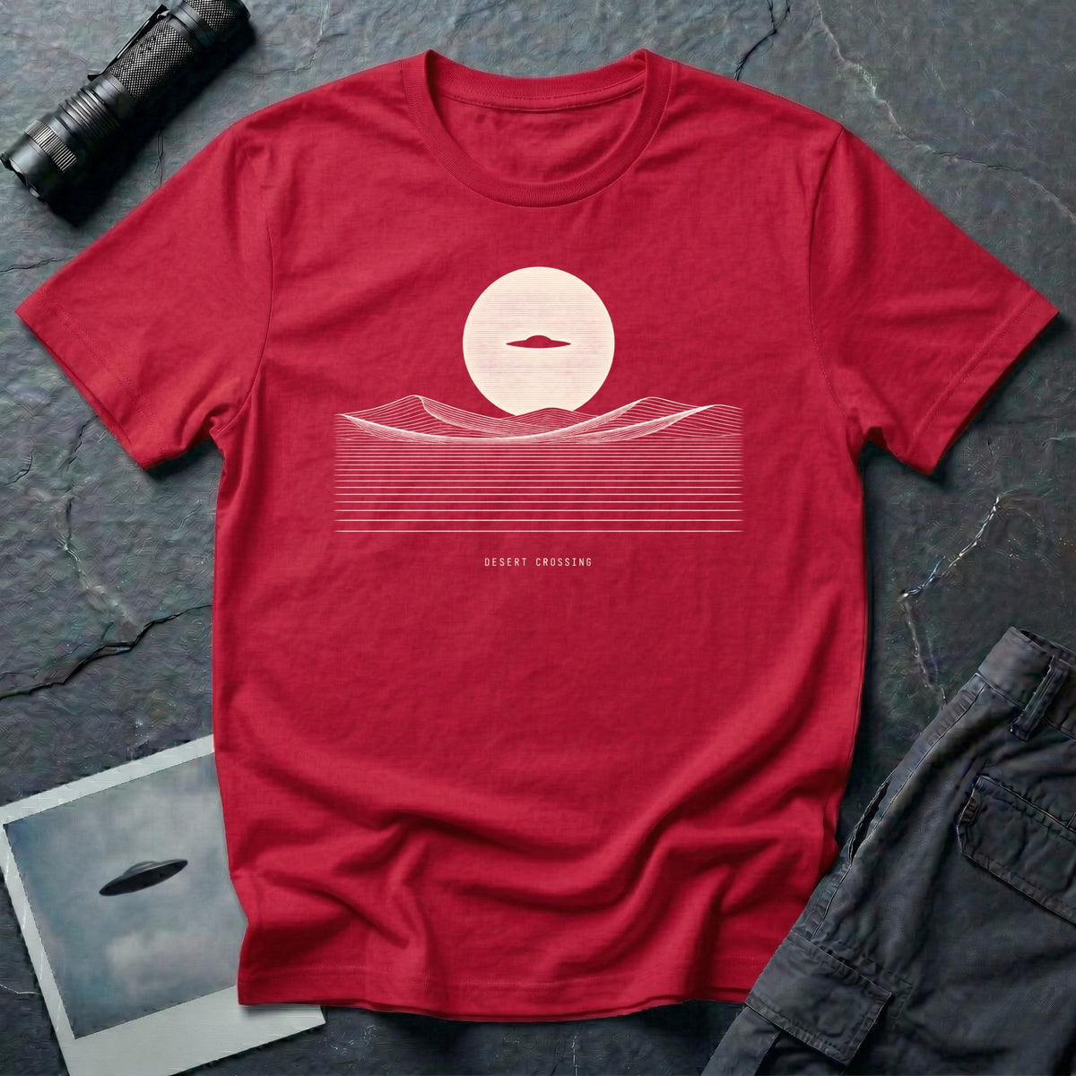 Desert Crossing UFO T-shirt with a black flying saucer crossing a white moon above minimalist line-art desert dunes, red tee