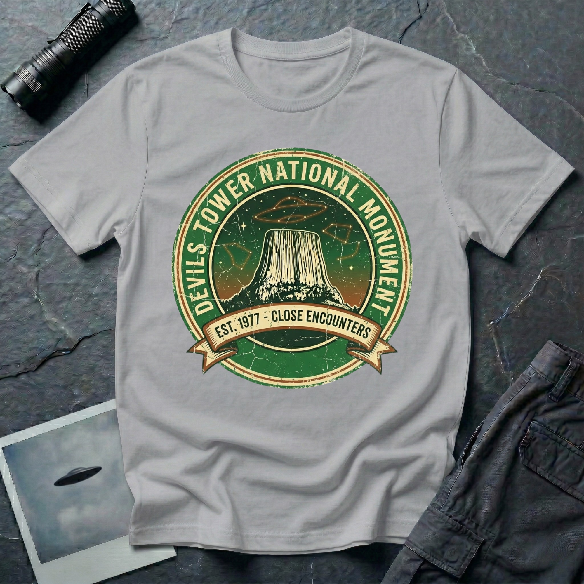 Devils Tower UFO t-shirt — vintage distressed National Park badge with the Wyoming monument silhouette, tiny flying saucers and constellations over a starry green sky, "DEVILS TOWER NATIONAL MONUMENT" arc text and "EST. 1906 — CLOSE ENCOUNTERS" banner on grey cotton tee