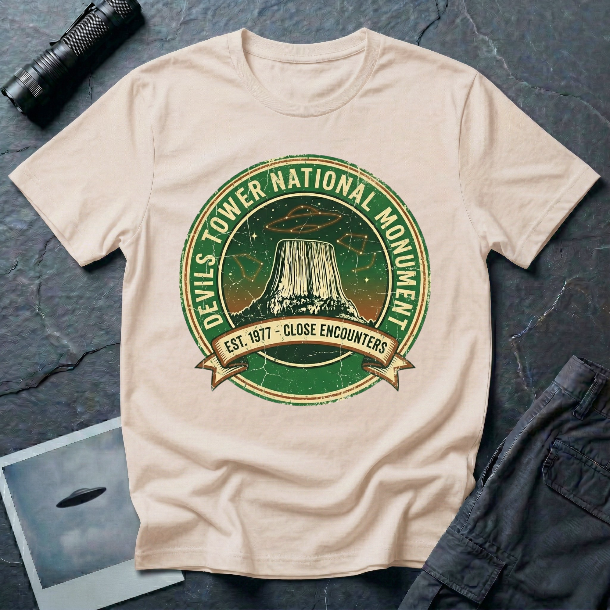 Devils Tower UFO t-shirt — vintage distressed National Park badge with the Wyoming monument silhouette, tiny flying saucers and constellations over a starry green sky, "DEVILS TOWER NATIONAL MONUMENT" arc text and "EST. 1906 — CLOSE ENCOUNTERS" banner on natural cotton tee