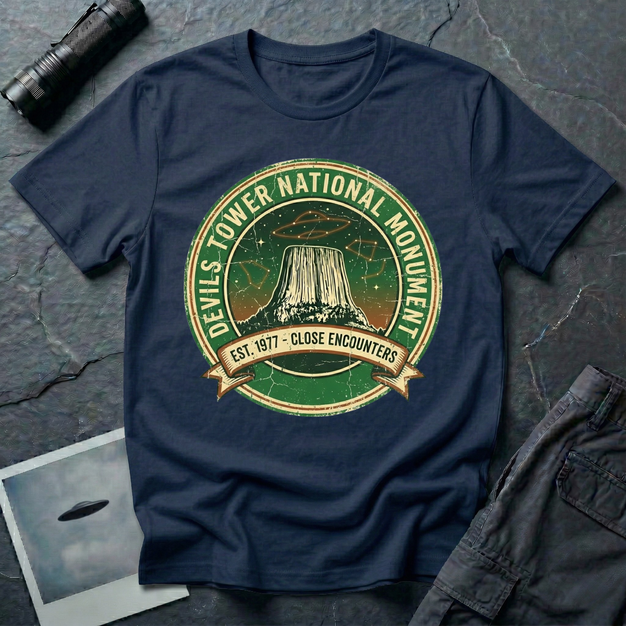 Devils Tower UFO t-shirt — vintage distressed National Park badge with the Wyoming monument silhouette, tiny flying saucers and constellations over a starry green sky, "DEVILS TOWER NATIONAL MONUMENT" arc text and "EST. 1906 — CLOSE ENCOUNTERS" banner on navy cotton tee