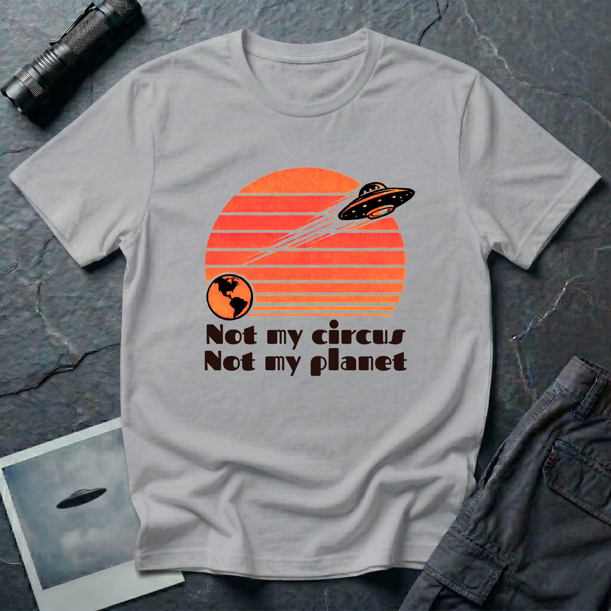 Not My Circus retro 1970s UFO t-shirt — flying saucer leaving Earth against striped sunset with "not my circus, not my planet" type on grey unisex cotton tee