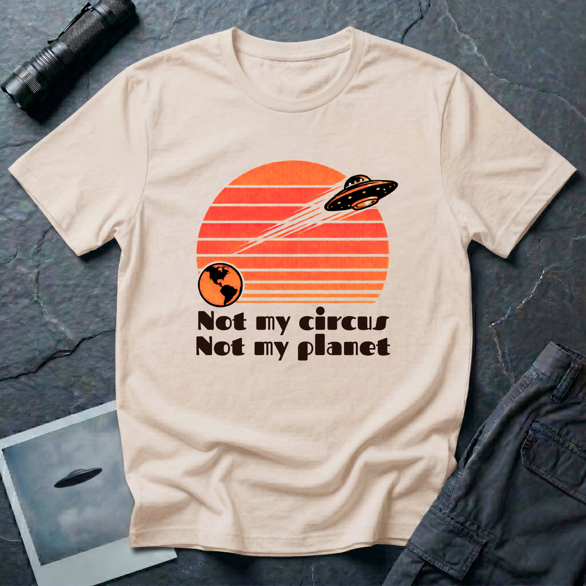Not My Circus retro 1970s UFO t-shirt — flying saucer leaving Earth against striped sunset with "not my circus, not my planet" type on natural unisex cotton tee