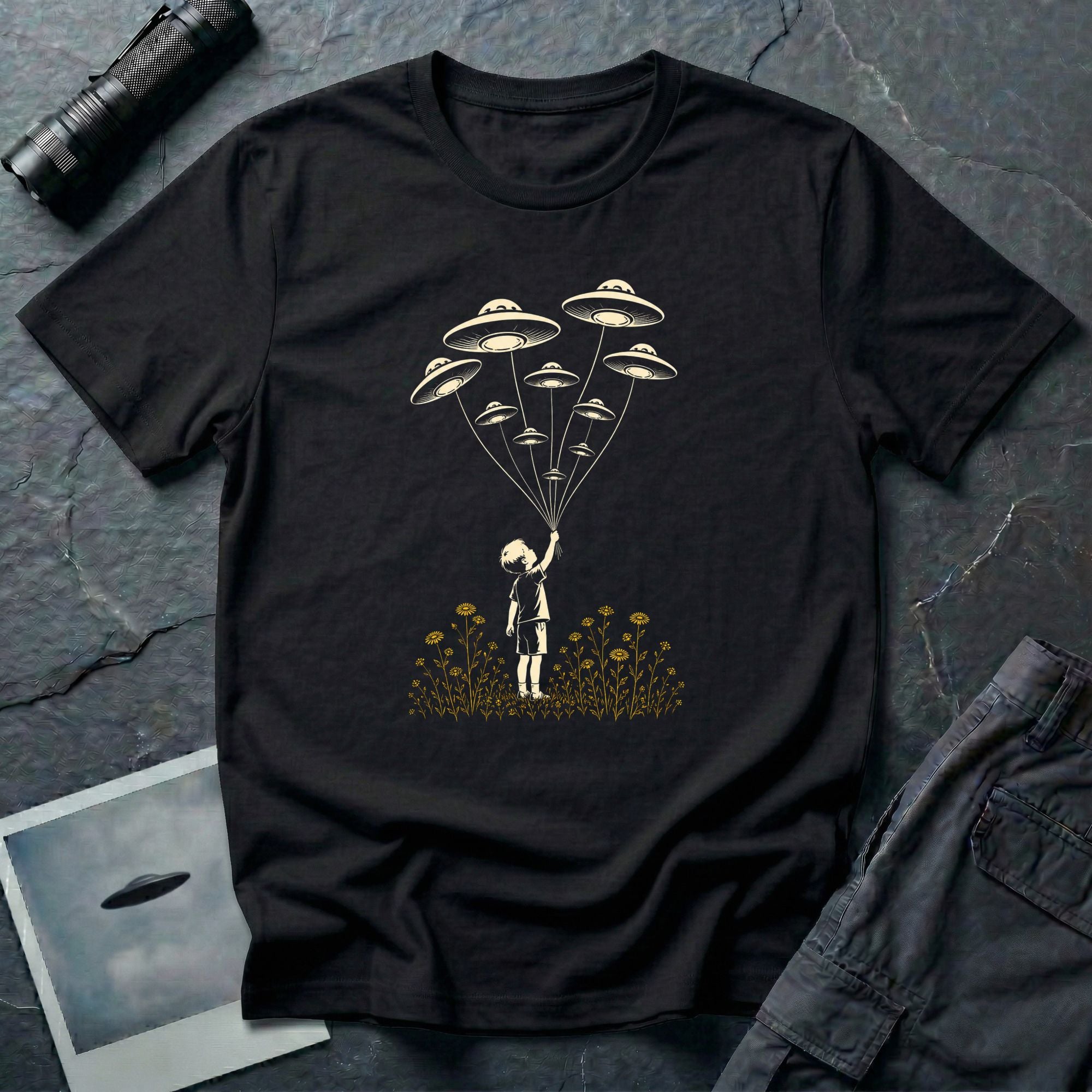Stencil UFO t-shirt design, child reaching up to hold strings tethered to a small fleet of UFOs, orange wildflowers at the base, black tee
