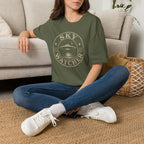 Person wearing green retro UFO t-shirt with a distressed circular badge showing a flying saucer over water at night with a crescent moon and the words SKY WATCHER in pale lettering