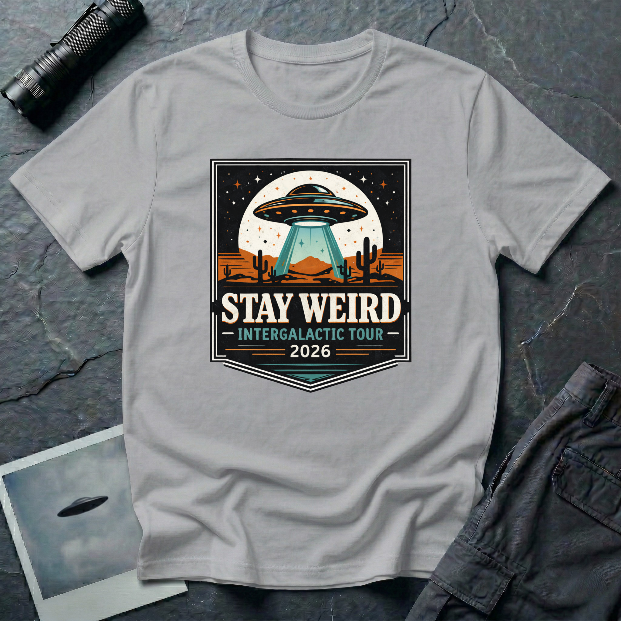Stay Weird desert UFO t-shirt — tour-poster badge with flying saucer over desert landscape and cacti on grey cotton tee