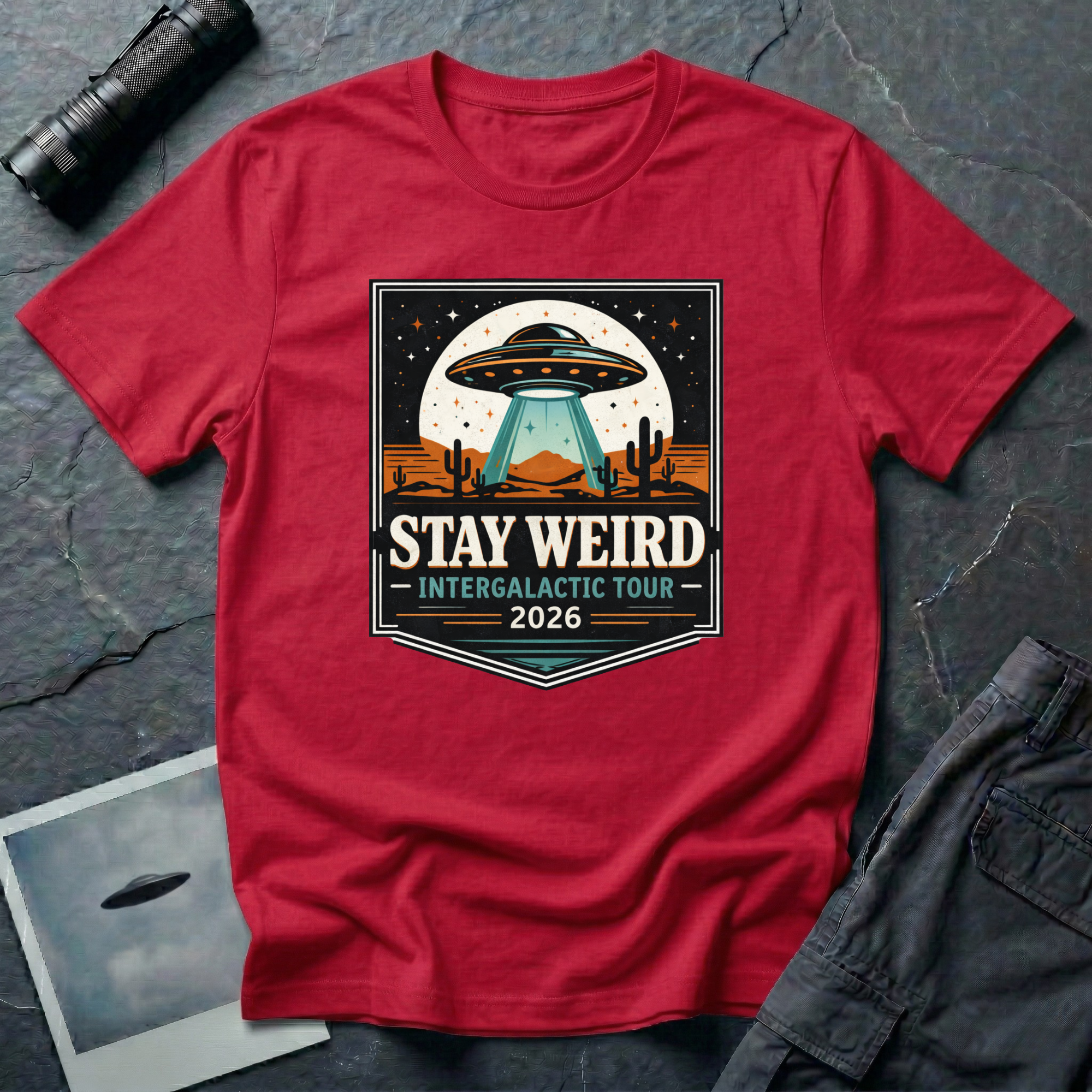 Stay Weird desert UFO t-shirt — tour-poster badge with flying saucer over desert landscape and cacti on red cotton tee