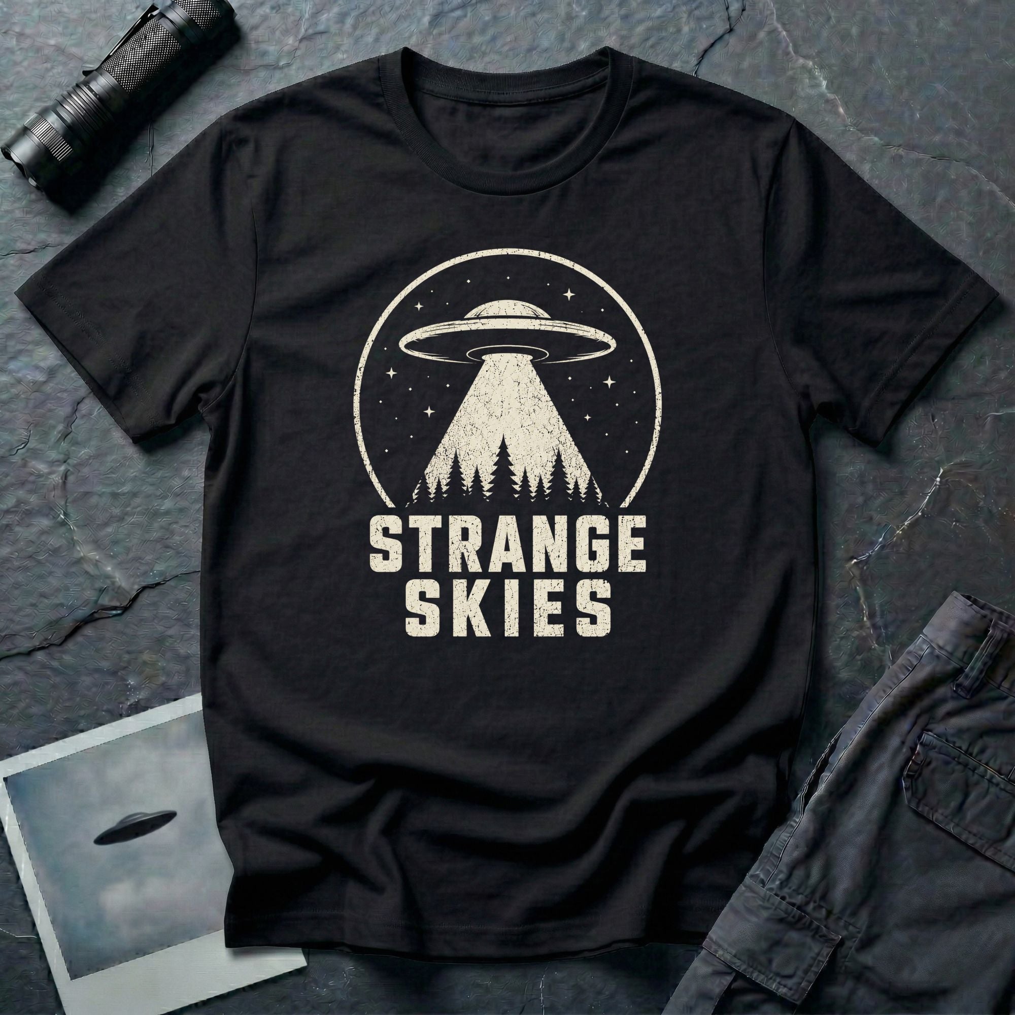 Black retro UFO t-shirt with a flying saucer and tractor beam over a conifer forest inside a distressed circle with the words STRANGE SKIES in white