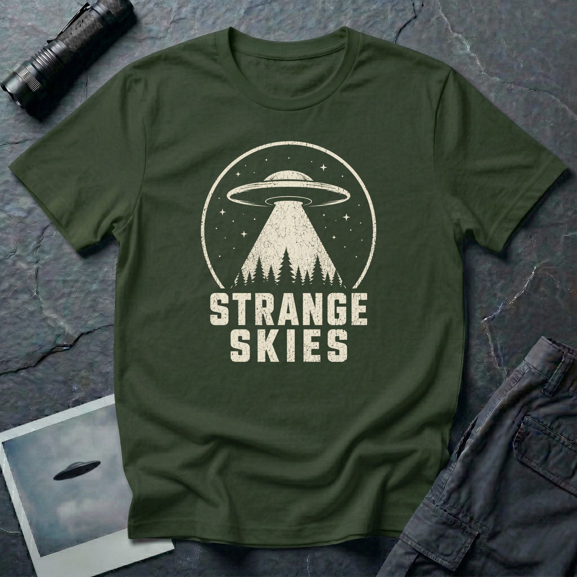 Military green retro UFO t-shirt with a flying saucer and tractor beam over a conifer forest inside a distressed circle with the words STRANGE SKIES in white