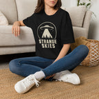 Person wearing retro UFO t-shirt with a flying saucer and tractor beam over a conifer forest inside a distressed circle with the words STRANGE SKIES in white
