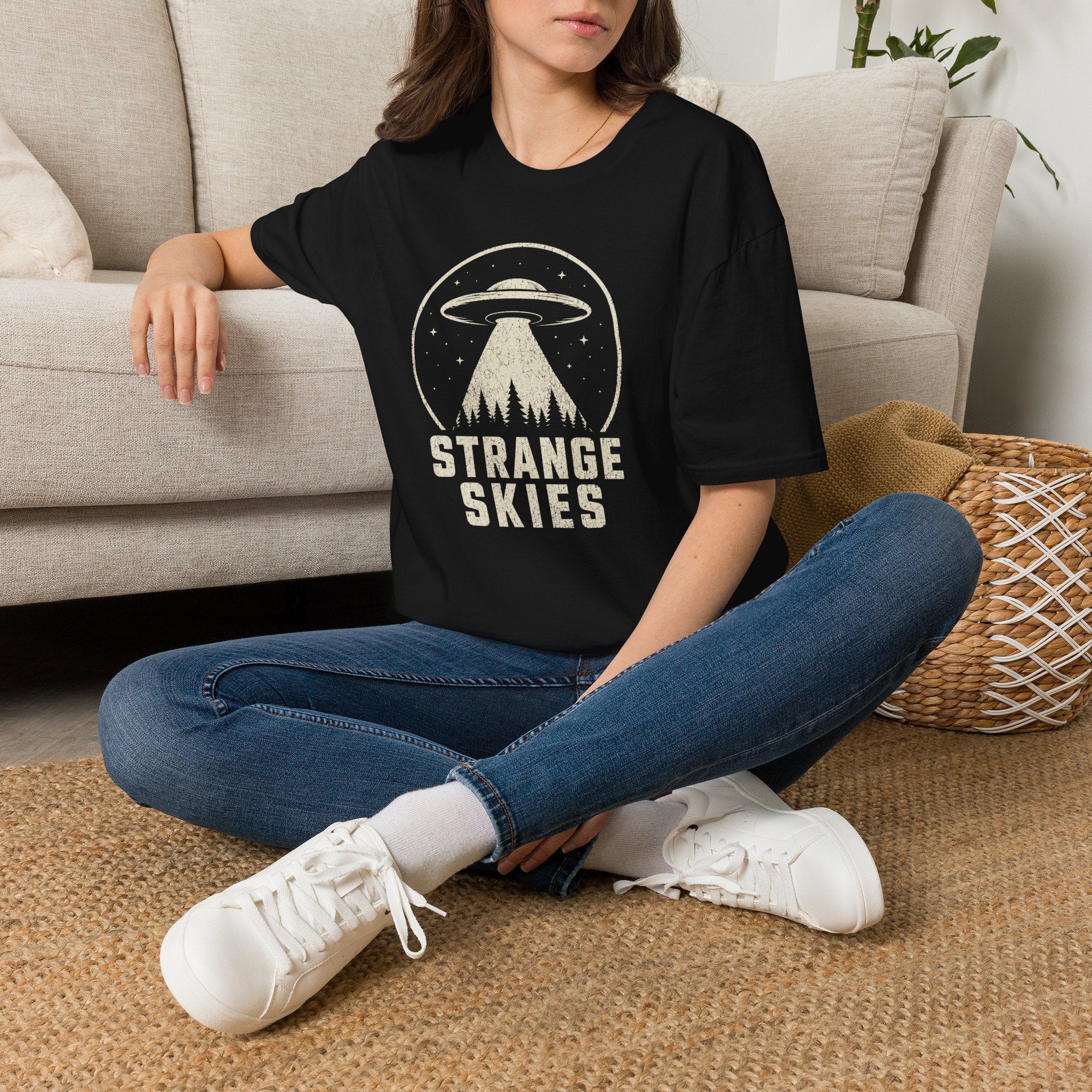 Person wearing retro UFO t-shirt with a flying saucer and tractor beam over a conifer forest inside a distressed circle with the words STRANGE SKIES in white