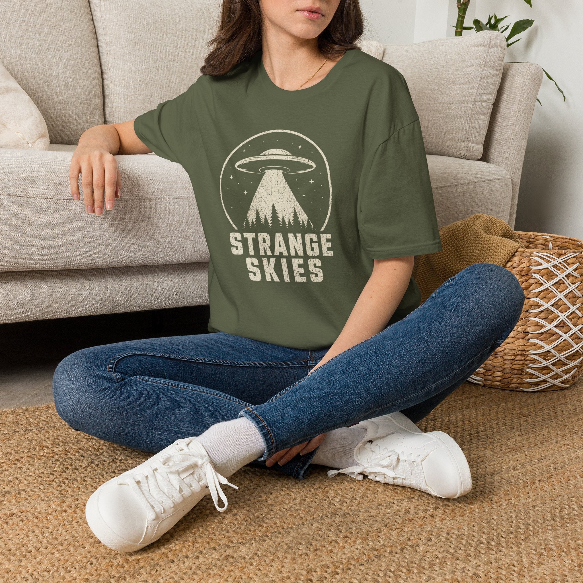 Person wearing retro UFO t-shirt with a flying saucer and tractor beam over a conifer forest inside a distressed circle with the words STRANGE SKIES in white
