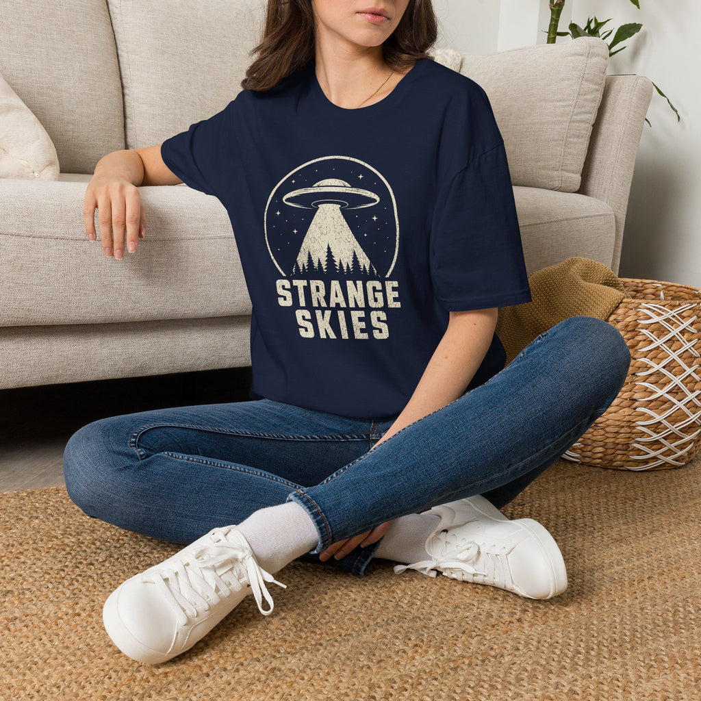 Person wearing navy blue retro UFO t-shirt with a flying saucer and tractor beam over a conifer forest inside a distressed circle with the words STRANGE SKIES in white