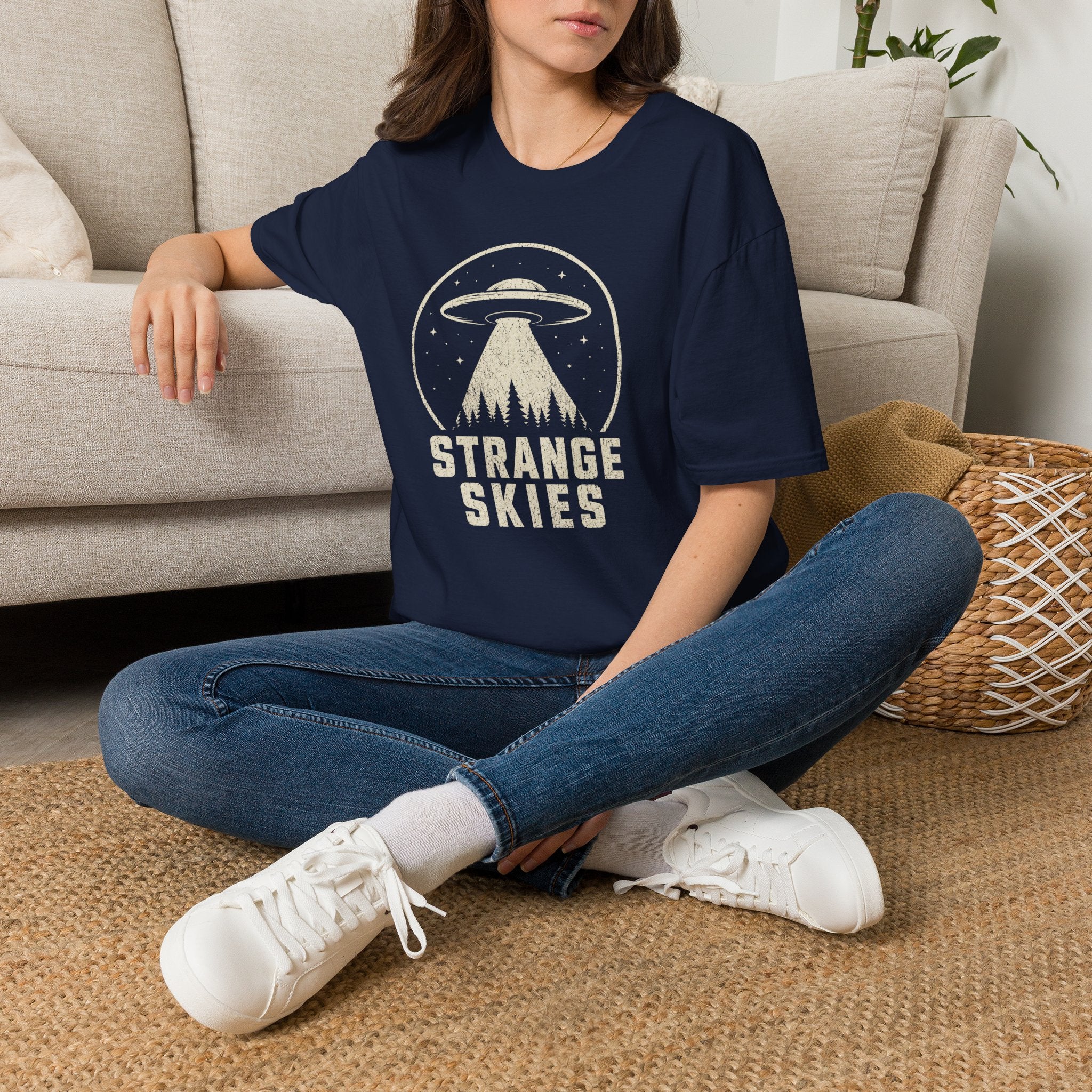 Person wearing navy blue retro UFO t-shirt with a flying saucer and tractor beam over a conifer forest inside a distressed circle with the words STRANGE SKIES in white