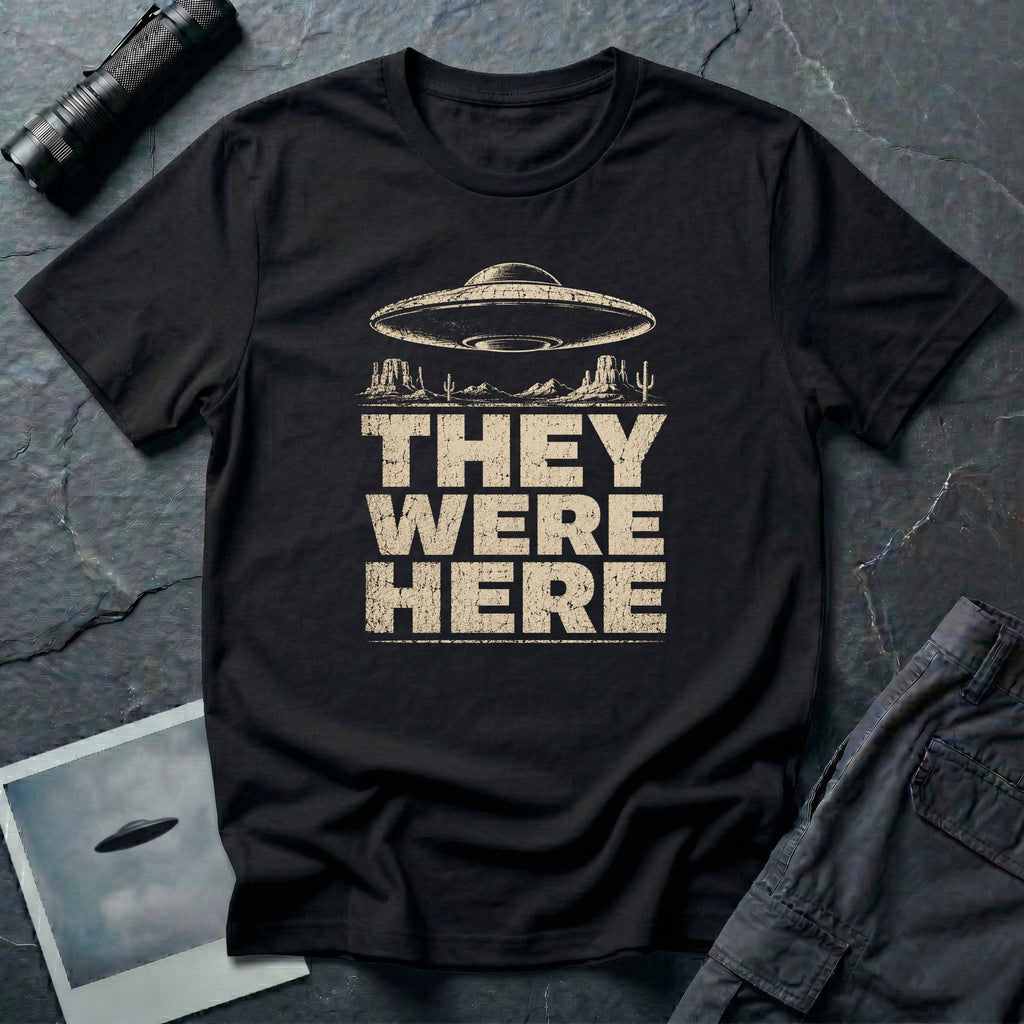 Black retro UFO t-shirt with a flying saucer over a southwest desert landscape with mesa formations and cacti and the words THEY WERE HERE in distressed pale block letters
