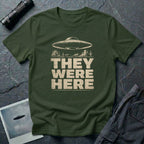 Green retro UFO t-shirt with a flying saucer over a southwest desert landscape with mesa formations and cacti and the words THEY WERE HERE in distressed pale block letters