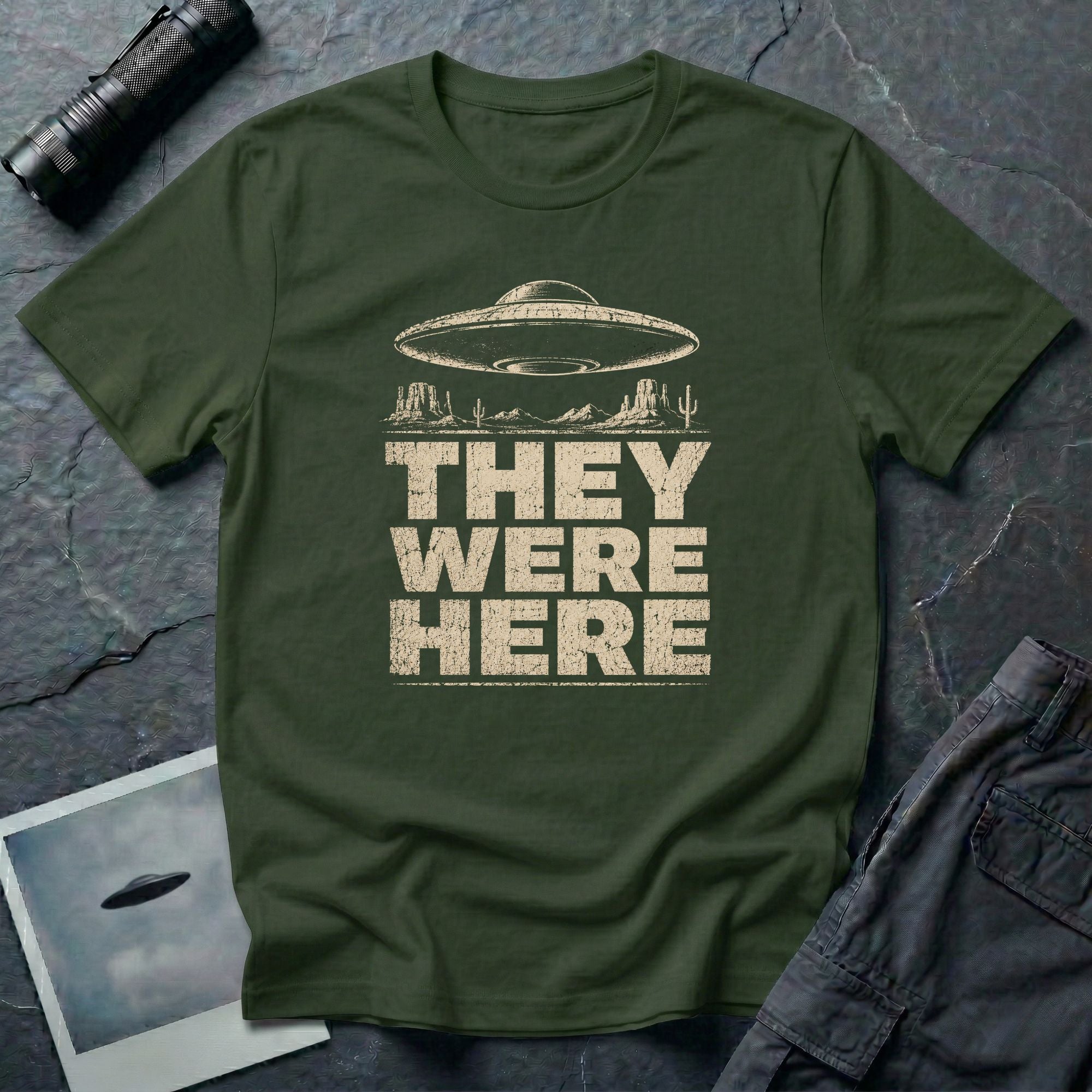 Green retro UFO t-shirt with a flying saucer over a southwest desert landscape with mesa formations and cacti and the words THEY WERE HERE in distressed pale block letters