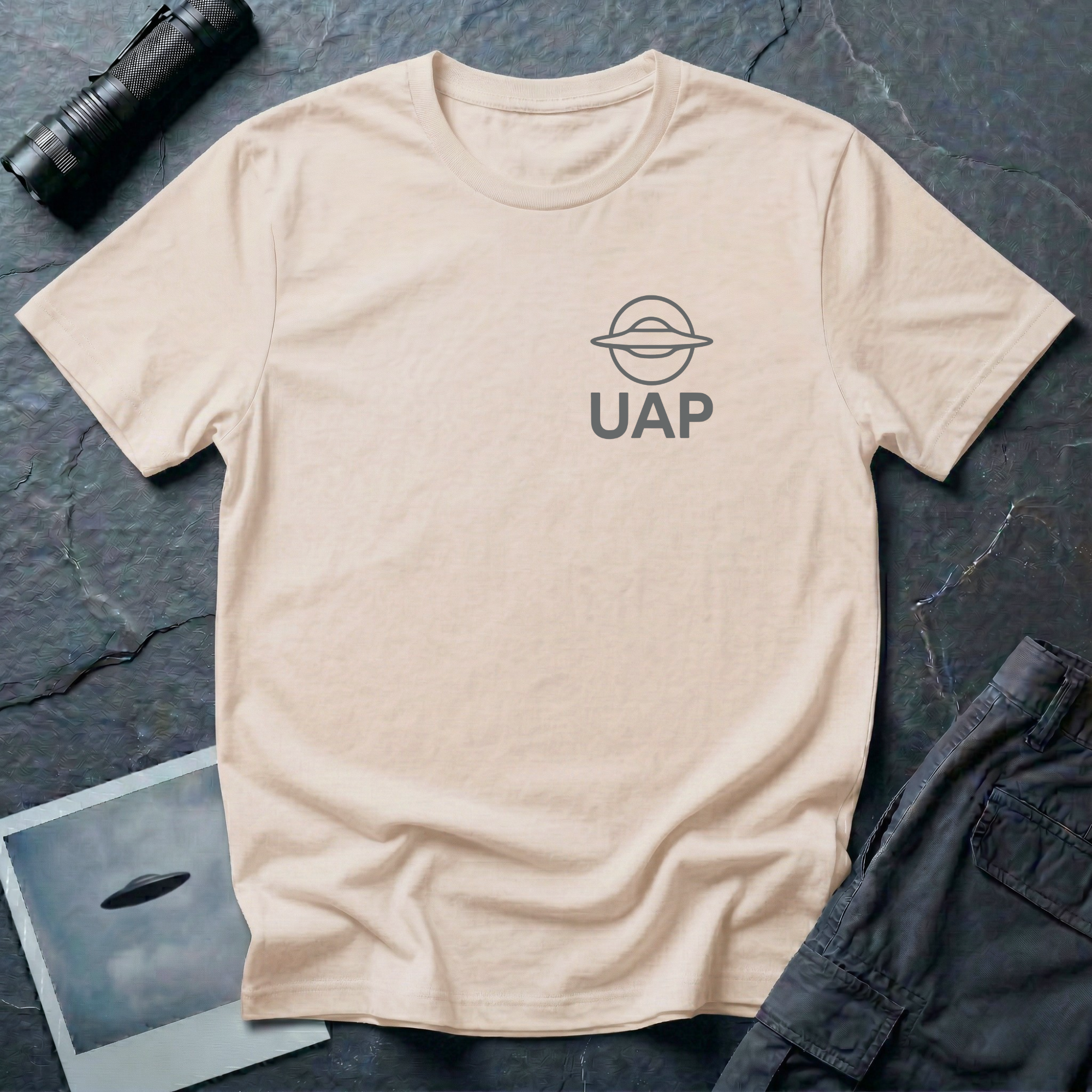 UAP logo t-shirt — small minimalist saucer icon and UAP letters in grey on the upper left chest of natural cotton tee