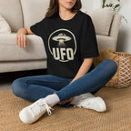Person wearing black retro UFO t-shirt with a distressed circle containing a flying saucer with tractor beam and bold block letters spelling UFO in white