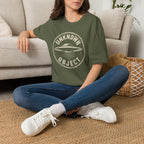 Person wearing green retro UFO t-shirt with a distressed circular badge showing a flying saucer with speed lines and the words UNKNOWN OBJECT in white