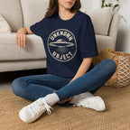 Person wearing navy blue retro UFO t-shirt with a distressed circular badge showing a flying saucer with speed lines and the words UNKNOWN OBJECT in white