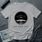 Grey t-shirt with a UFO hovering over mountains and a reflective lake inside a circular night scene badge with the words WE SEE YOU in black and gold