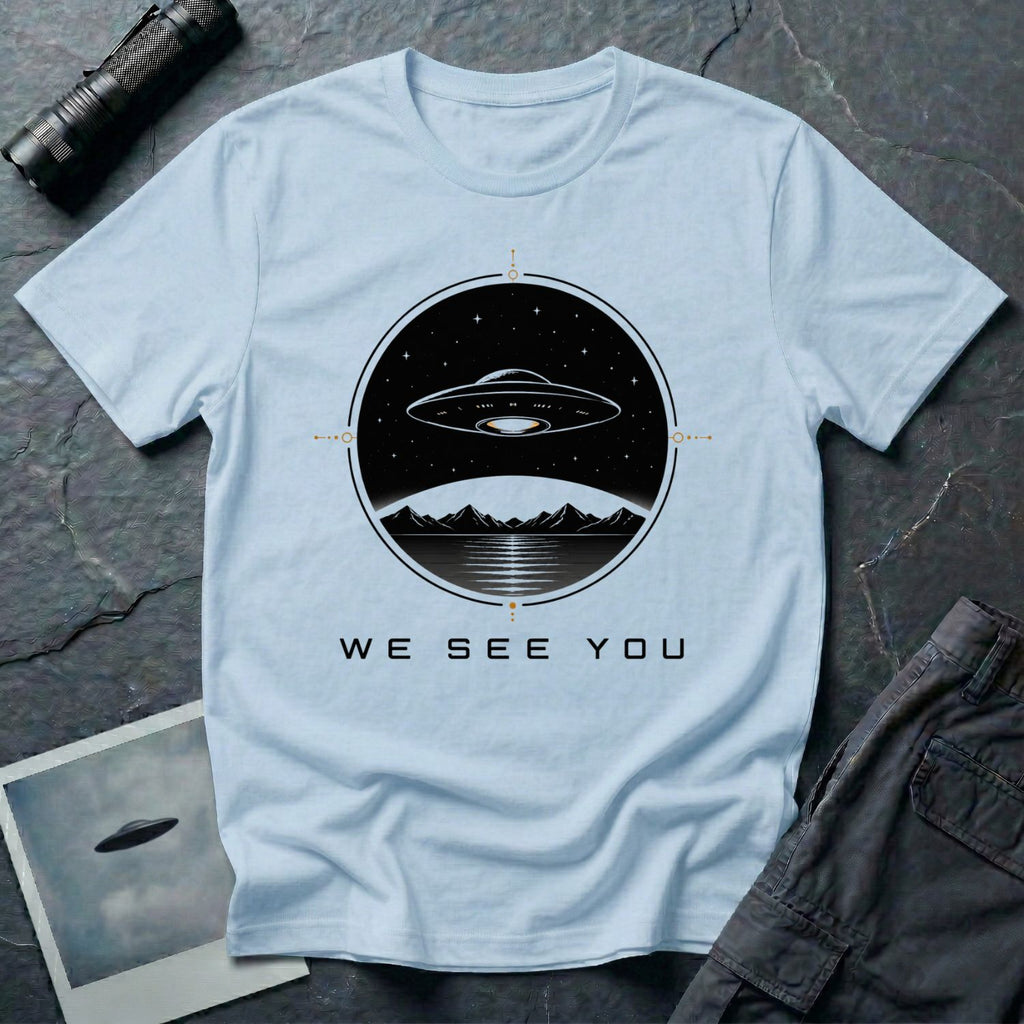Light blue t-shirt with a UFO hovering over mountains and a reflective lake inside a circular night scene badge with the words WE SEE YOU in black and gold