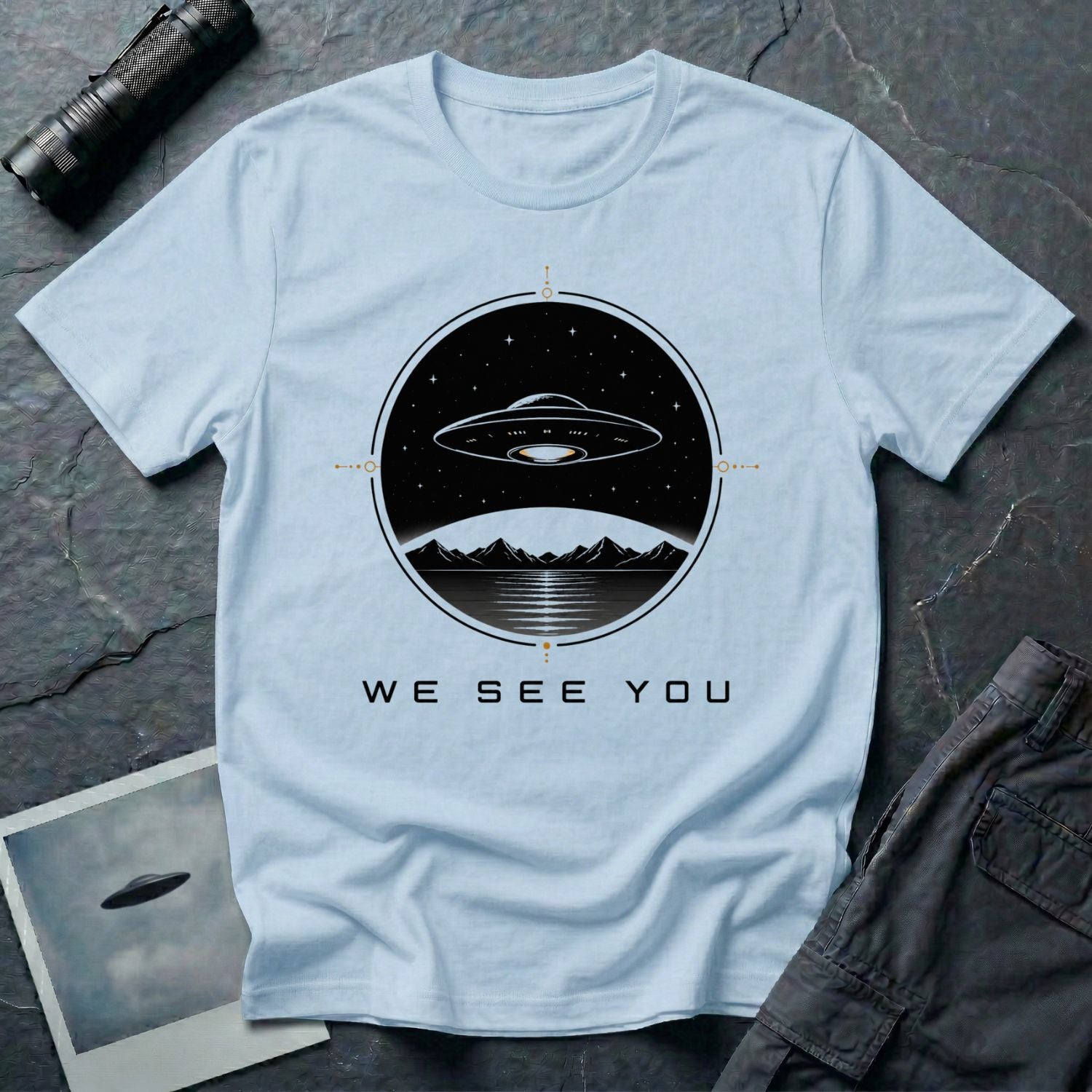 Light blue t-shirt with a UFO hovering over mountains and a reflective lake inside a circular night scene badge with the words WE SEE YOU in black and gold