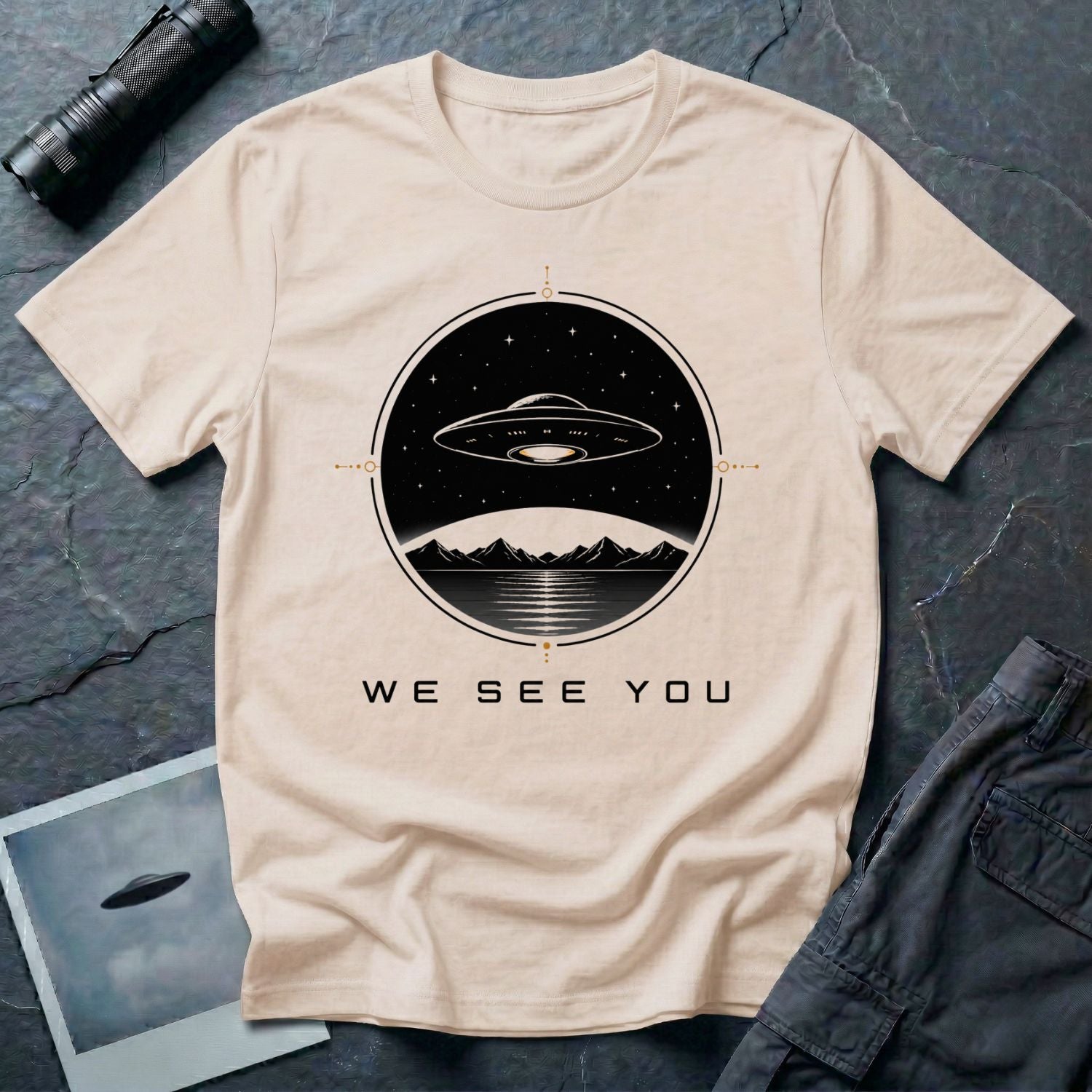Natural t-shirt with a UFO hovering over mountains and a reflective lake inside a circular night scene badge with the words WE SEE YOU in black and gold