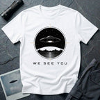 White t-shirt with a UFO hovering over mountains and a reflective lake inside a circular night scene badge with the words WE SEE YOU in black and gold