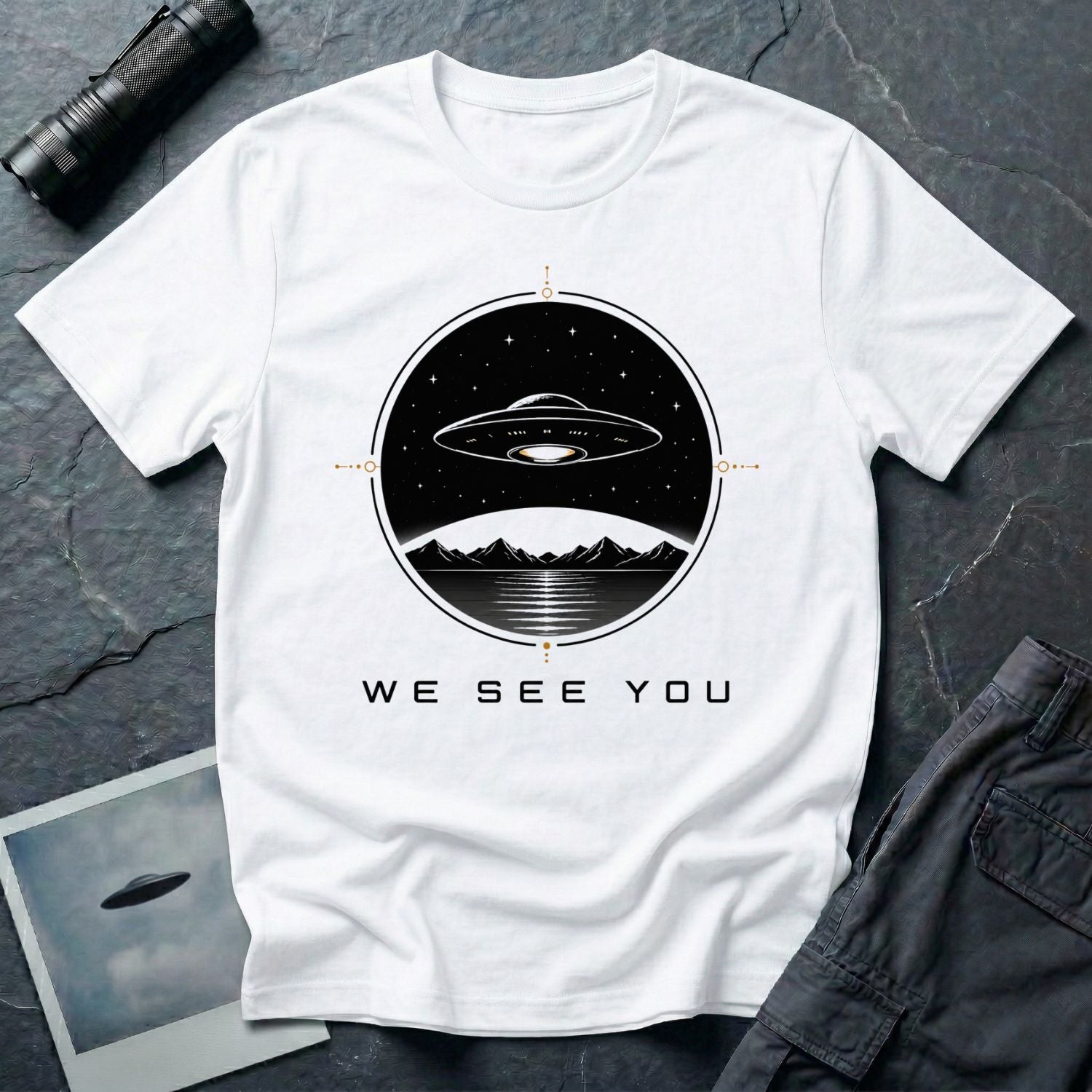 White t-shirt with a UFO hovering over mountains and a reflective lake inside a circular night scene badge with the words WE SEE YOU in black and gold